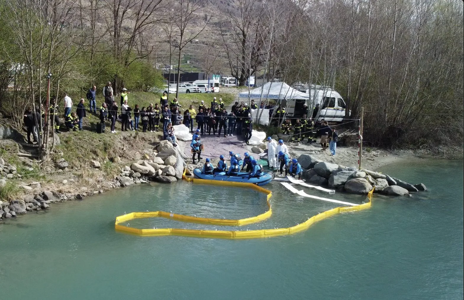 Yellow containment boom curved on a river with a boat and response teams on the rocky bank