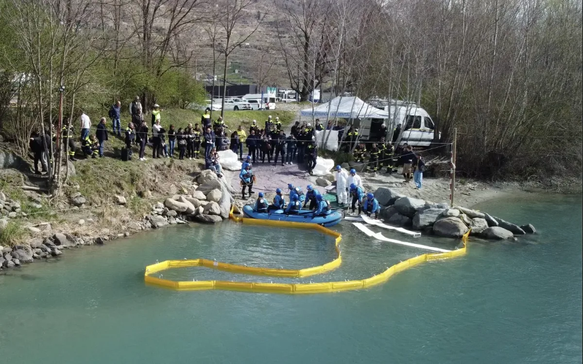 Yellow containment boom curved on a river with a boat and response teams on the rocky bank
